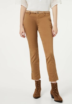 Person trägt hellbraune Slim-Fit-Hose mit einem dünnen Gürtel und braunen Wildleder-Stiefeletten, die mit silbernen Sternen verziert sind.