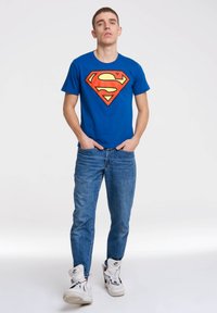 Blue cotton t-shirt featuring a large red and yellow Superman logo on the front, paired with light blue jeans and white sneakers.