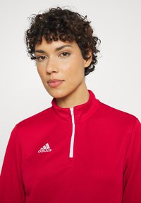 Rotes Sportoberteil mit Stehkragen und weißem Reißverschluss. Verfügt über ein Adidas-Logo auf der linken Brustseite. Glatter, strukturierter Stoff.