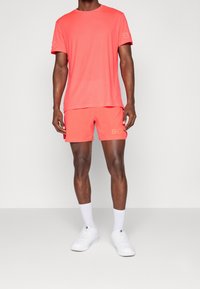 Korálový sportovní set trička a šortek, vyrobený z lehkého materiálu, s kulatým výstřihem, krátkými rukávy a jemným logem na šortkách.