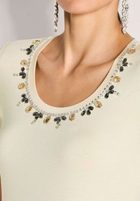 Close-up van de halslijn van een crèmekleurige top, versierd met zwarte, amberkleurige en heldere rhinestones, gedragen met grote hangende rhinestone oorbellen.