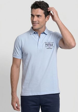 Hombre con cabello oscuro que lleva una camiseta polo azul claro con texto en el pecho, tocándose el pelo y mirando hacia un lado, de pie contra un fondo liso.