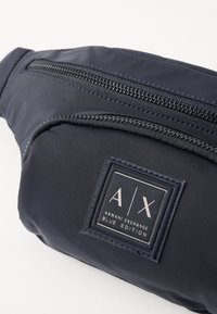 Sac banane en tissu bleu marine avec plusieurs compartiments zippés, doté d'un patch logo rectangulaire étiqueté "A|X ARMENI EXCHANGE BLUE EDITION."