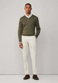 Olive green v-neck sweater, white collared shirt, light cream trousers, and brown lace-up shoes, standing against a neutral background.