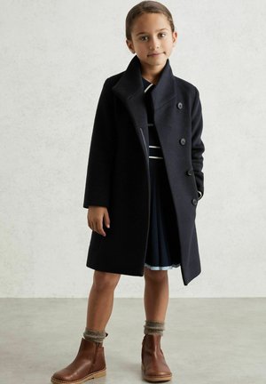 Cappotto di lana navy con ampio colletto e cinque bottoni, indossato sopra un vestito a righe. Il modello indossa stivaletti marroni alla caviglia e calze grigie lavorate a maglia.