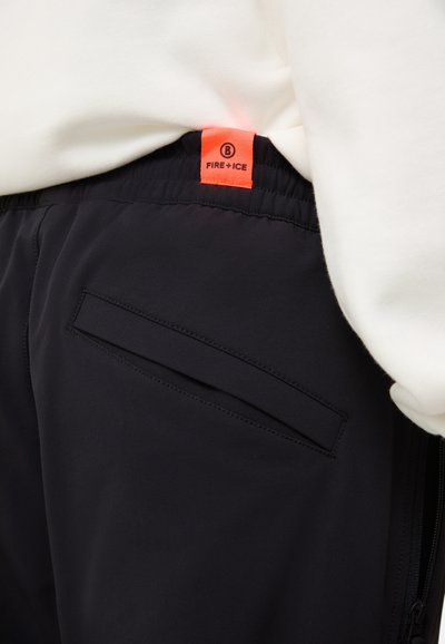 Sorte syntetiske bukser med elastisk talje, der har et orange label med teksten "FIRE + ICE" og en flad baglomme med en diskret søm.