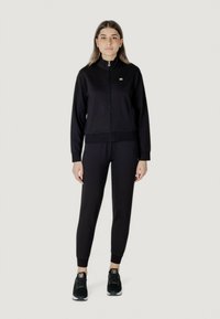 Tuta nera con giacca a zip e pantaloni aderenti, realizzata in tessuto liscio. Presenta un colletto alto e polsini elasticizzati. Sneakers nere completano il look.