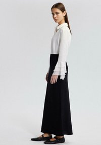 Blusa blanca con mangas acampanadas combinada con una larga falda negra. El conjunto presenta un diseño de cuello y zapatos negros planos.
