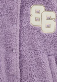 Lilla fliisjope tekstuurse viimistlusega. Suur valge "86" tikand kuldse äärega esiküljel. Esiküljel nööpkinnitused.