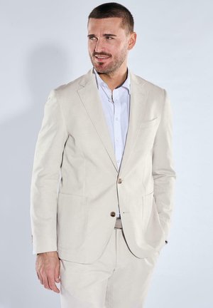 Mann mit kurzen Haaren und Bart, der einen cremefarbenen Anzug und ein hellblaues Hemd trägt, steht mit einer Hand in der Tasche und schaut nach rechts.