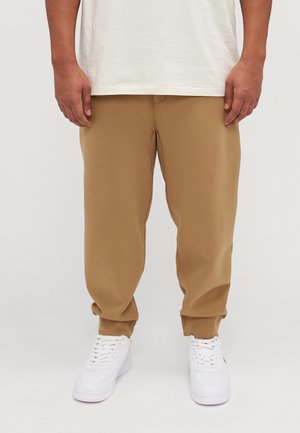 Osoba ubrana w beżowe spodnie dresowe i białe trampki, stojąca z rękami swobodnie opuszczonymi wzdłuż ciała na neutralnym tle.