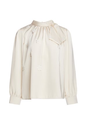 Blouse crème à manches longues avec un col montant noué en nœud et de petites applications florales éparpillées sur le tissu.