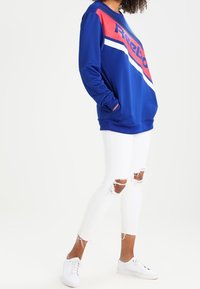 Blå sweatshirt med röda accenter och vita ränder, med en framträdande Reebok-logotyp; kombinerad med vita rivna skinny jeans och sneakers.