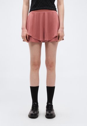 Personne portant une jupe à volants rose poudré, des chaussettes noires et des chaussures noires à lacets, debout devant un fond blanc uni.