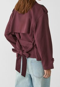 Bordeaux gekorte jas met achterknoopjes, schouderepauletten en een gladde textuur, gecombineerd met lichtblauwe jeans met details van de achterzakken.
