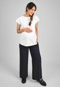 Weißes kurzärmliges Oberteil mit abgerundetem Saum, kombiniert mit locker sitzenden marineblauen Hosen. Das Model steht mit einer Hand auf dem Bauch und präsentiert eine entspannte Passform.