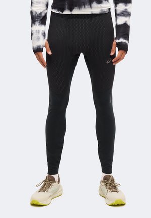 Persona che indossa leggings sportivi neri texturizzati, scarpe da corsa bianche e marroni con suola gialla e una maglia tie-dye bianco e nero.