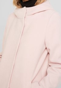 Cappotto di lana rosa chiaro con colletto alto, maniche lunghe e un design minimalista, dotato di tasche laterali e una texture morbida.