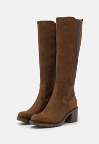 Bottes hauteur genou en daim marron avec panneaux latéraux élastiques et talon bloc. Présentent une texture lisse et une fermeture éclair sur le côté intérieur.