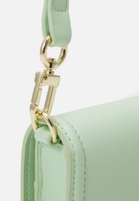 Ljust grön läderväska med hörn, guldmetallklämma och ring som fäster en matchande grön rem, visad mot en vit bakgrund.
