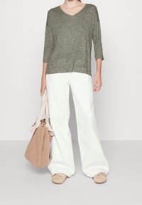 Pull en laine olive à col en V avec manches trois-quarts, associé à un pantalon blanc ample. Le mannequin tient un sac en tissu beige et porte des chaussures beige clair.