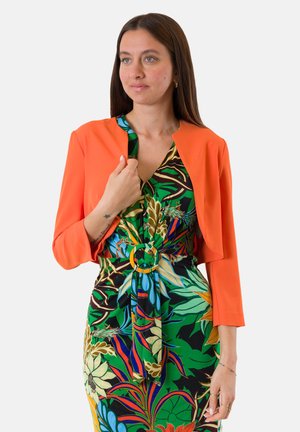 Donna con un vivace abito floreale con fibbia rotonda alla cintura indossa una giacca corta arancione brillante, guardando leggermente di lato.