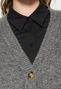 Grijze gebreide cardigan met een V-hals, met een gestructureerde afwerking en een bruine knoopsluiting. Gedragen over een zwart shirt met kraag.