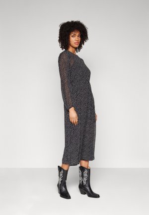 Schwarzes Midikleid mit weißen Punkten, langen transparenten Ärmeln und lässiger Passform. Kombiniert mit schwarzen Cowboystiefeln mit weißen Details.