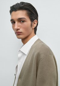 Beige gestrickte Strickjacke über einem weiß gestreiften Hemd, mit lässigem Schnitt und subtiler Textur. Neutrale, gedämpfte Farben dominieren das Outfit.