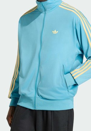 Mann trägt eine hellblaue Adidas Trainingsjacke mit gelben Streifen an den Ärmeln und Logo, Hand in der Tasche, kombiniert mit schwarzen Hosen.