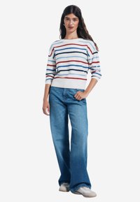 Gestreifter Strickpullover mit blauen, roten und weißen Linien; breite Bündchen und Saum. Kombiniert mit hoch geschnittenen, ausgestellten blauen Jeans und hellen Sneakers.