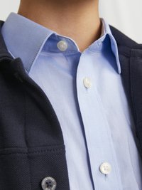 Veste navy avec une chemise à col bleu visible. Les boutons sont blancs, et la chemise a une texture lisse. La veste a un tissu texturé.