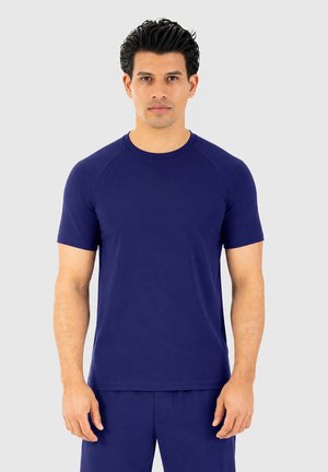 Hombre con cabello oscuro que lleva una camisa de manga corta azul oscuro lisa y pantalones a juego, de pie frente a un fondo claro y liso.