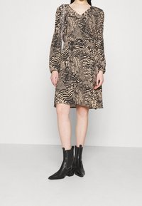 Femme portant une robe imprimée animal beige et noire à manches longues, ainsi que des bottines noires à talons bloc.