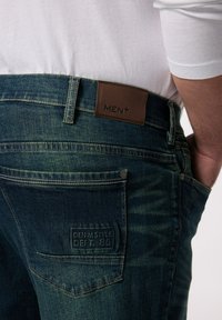 Džíny z denimu s tmavě modro-zeleným omakem, vyznačující se hnědou koženou nášivkou s nápisem "MEN+" a nášivkou na zadní kapse označenou "DENIM STYLE DEPT. 36".