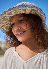 Cappello di paglia intrecciato con dettagli blu, crema e verde; texture morbida; indossato con una maglietta leggera color crema con scollo a V; sfondo soleggiato.