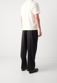 Lichtbeige kortarmshirt in combinatie met loszittende zwarte broek en zwarte schoenen, gezien van achteren tegen een witte achtergrond.