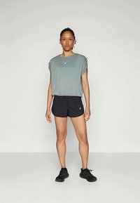 En kvinne står med ansiktet vendt fremover iført en løs grå treningsgenser, svarte shorts og svarte joggesko foran en lys, ensfarget bakgrunn.