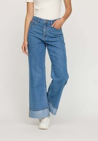 Jean bleu en denim à jambes larges avec une taille haute, des accents d'ourlet effiloché et un délavage clair, porté avec un haut blanc et des baskets blanches.
