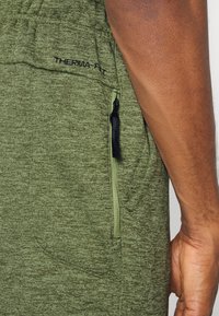 Calções Therma-FIT verdes com um tecido texturizado, apresentando um bolso lateral com fecho e um logótipo bordado na faixa da cintura.