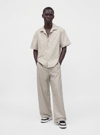 Homme debout, mains dans les poches, portant une chemise beige à manches courtes à motifs et un pantalon large assorti avec des baskets noires et blanches.