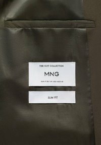 Das Innere der Anzugjacke zeigt zwei weiße Etiketten mit "THE SUIT COLLECTION MNG" und "SLIM FIT", die auf olivgrünem Stoff gedruckt sind.