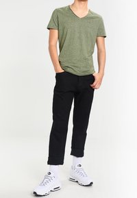 T-shirt verde a scollo a V, pantaloni neri con risvolto e sneakers bianche con accenti neri. La t-shirt presenta un piccolo logo sul petto.