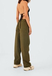 Vrouw in olijfgroene cargo pants, beige sneakers en een zwarte haltertop zonder rug met gestrikte banden, staande tegen een effen lichte achtergrond.