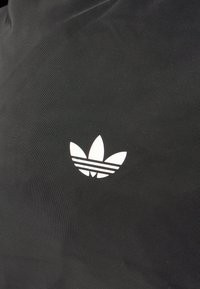 Μαύρο ύφασμα με λευκό λογότυπο Adidas trefoil και τρεις οριζόντιες ρίγες. Η υφή είναι λεία και ματ.