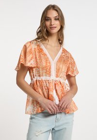 Blusa arancione a fantasia con maniche a campana corte, scollo a V, rifiniture in pizzo e vita regolabile con dettaglio a nappina. Indossata con jeans chiari.