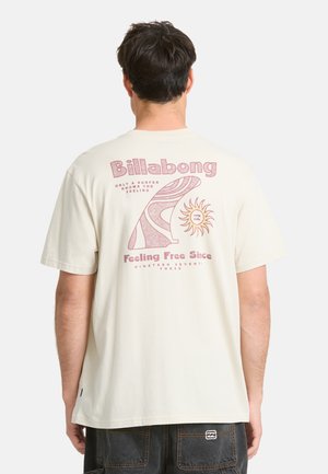 Mann trägt ein cremefarbenes T-Shirt mit pinkem Billabong-Logo, stilisiertem Surfbrett- und Sonnengrafik sowie Text auf dem Rücken.