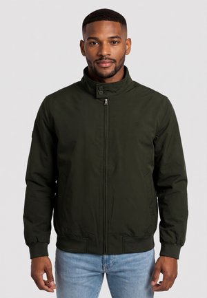 Uomo che indossa una giacca verde scuro con cerniera e collo con bottoni, e jeans azzurri, in piedi davanti a uno sfondo semplice.