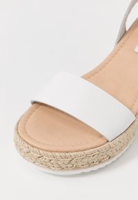 Sandale avec bride synthétique blanche, semelle intérieure rembourrée beige et plateforme en jute tressé. Présente une texture lisse et un design de semelle plate.