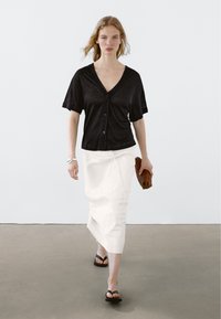 Svart kortärmad skjorta med knappar, vit texturerad kjol, svarta flip-flops, hållande brun vävd clutch, modell som går på en neutral golv.